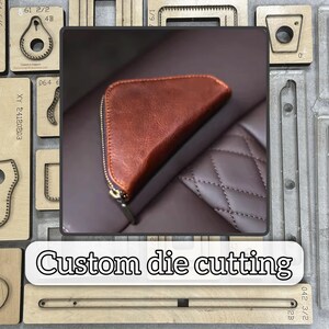 May include: A brown leather wallet with a zipper closure, resting on a brown surface. The wallet has a curved shape and a gold-colored zipper. The image also shows wooden die-cutting tools and the text "Custom die cutting".