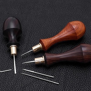 May include: A collection of leatherworking tools, including three awls with wooden handles in varying shades of brown and a metal L-shaped wrench. The awls have brass ferrules and sharp metal tips. Two spare needles are also present.