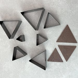 May include: A set of nine metal triangle-shaped cookie cutters in various sizes, arranged on a white textured surface. Three brown leather triangles are also included.