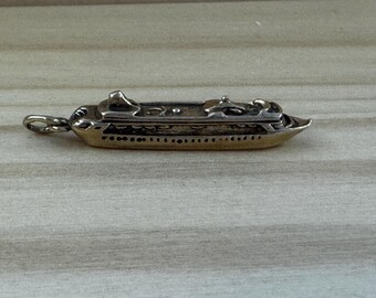May include: A gold-toned cruise ship pendant with silver accents. The miniature ship features detailed elements like smokestacks and windows. The pendant has a loop for attaching to a chain or bracelet. The background is a light wood grain.