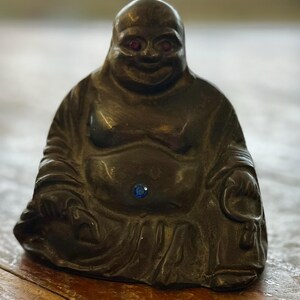 May include: A dark brown Buddha statue with red eyes and a blue gem on the belly. The statue is seated with a serene expression, and the details of the robes are visible. The statue is on a wooden surface.