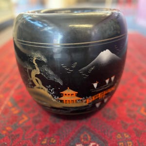 May include: Black ceramic jar with a Japanese-inspired design. The jar features a white mountain, a black tree, and a red and orange building with a gold roof.