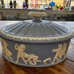 May include: A blue ceramic serving dish with a lid. The dish features a white floral handle and a decorative border. The sides are adorned with white relief figures. A classic piece for serving food.