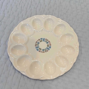 May include: A white ceramic egg serving platter with 12 egg-shaped indentations. The center features a small, colorful geometric design in blue, brown, and teal. The platter is round and has a scalloped edge.