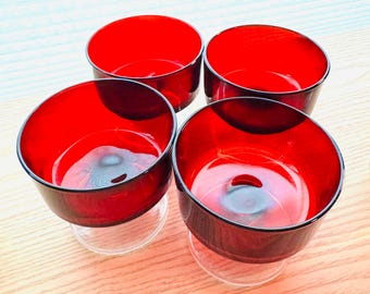 Könnte beinhalten: Vier rote Glasschalen mit klaren Glasbasen sind auf einer hellen Holzoberfläche angeordnet. Die Schalen haben einen tiefroten Innenraum und einen dunklen Rand. Die klaren Basen bieten Stabilität und einen Kontrast.