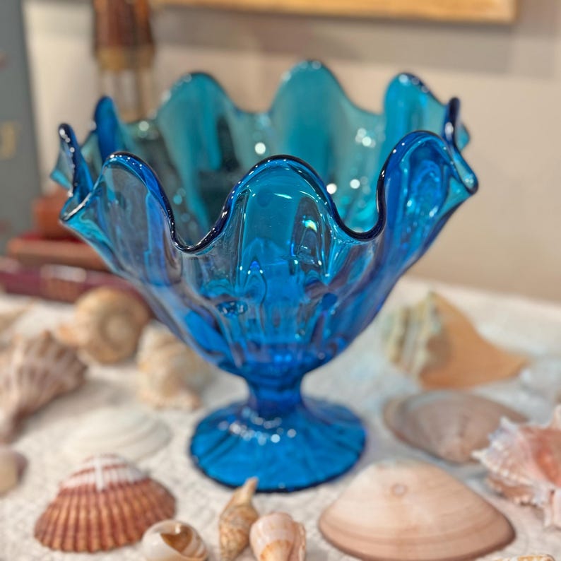 K&ouml;nnte beinhalten: Eine leuchtend blaue Glasschale mit gewelltem Rand, die auf einem Sockel steht. Die Schale ist von verschiedenen Muscheln umgeben, was ein K&uuml;stenmotiv erzeugt. Die Farbe der Schale ist ein tiefes, transparentes Blau.