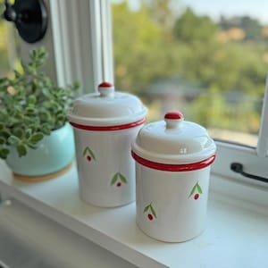 May include: Two white ceramic canisters with red accents and cherry designs. Each canister has a red knob on the lid and a red band around the top. Green leaves and red cherries are painted on the sides.