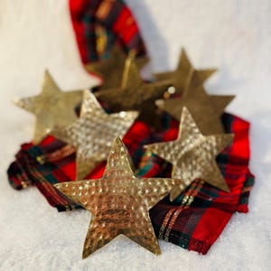 May include: Six hammered brass star ornaments rest on a red and green plaid fabric, set against a snowy white background. The stars have a textured surface and a warm, metallic finish, perfect for holiday decor.
