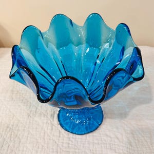 K&ouml;nnte beinhalten: Eine leuchtend t&uuml;rkisfarbene Glasschale mit ger&uuml;schtem Rand und Sockel. Die wellenf&ouml;rmigen Kanten der Schale sind schwarz umrandet, was einen auff&auml;lligen Kontrast erzeugt. Dieses dekorative St&uuml;ck ist ideal zum Servieren oder Ausstellen.