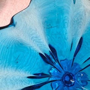 K&ouml;nnte beinhalten: Nahaufnahme einer dekorativen Glasschale mit blumenartigem Design. Die Schale weist Blaut&ouml;ne von hellem T&uuml;rkis bis tiefem Kobalt auf. Das Glas hat ein strukturiertes, wellenf&ouml;rmiges Aussehen mit einem zentralen, dunkleren blauen Blumenelement.