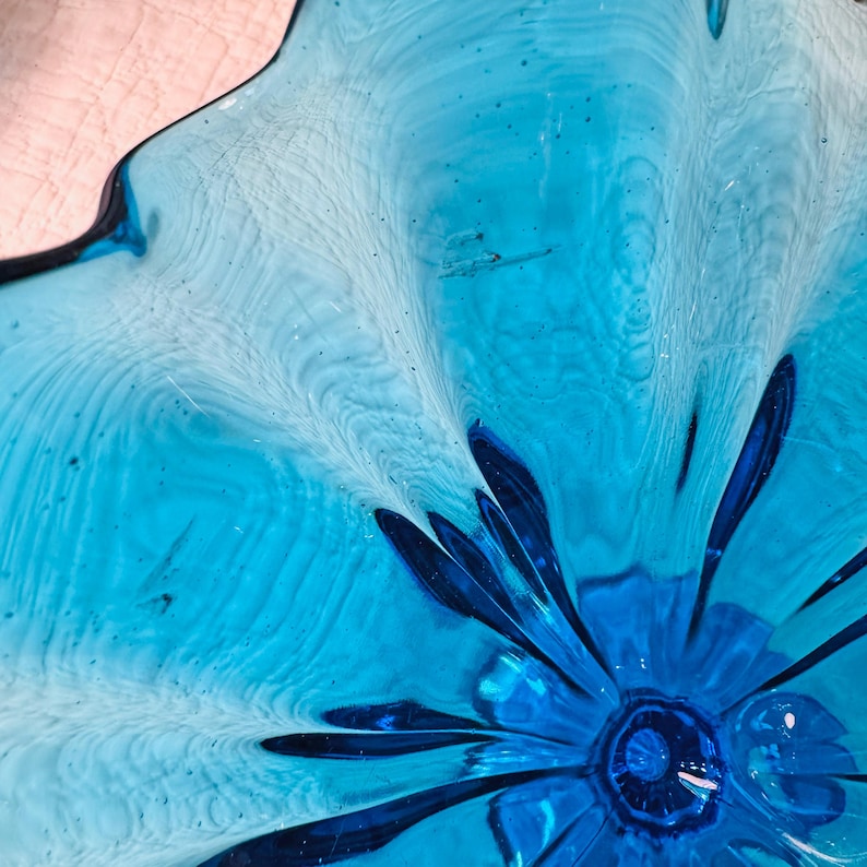 K&ouml;nnte beinhalten: Nahaufnahme einer dekorativen Glasschale mit blumenartigem Design. Die Schale weist Blaut&ouml;ne von hellem T&uuml;rkis bis tiefem Kobalt auf. Das Glas hat ein strukturiertes, wellenf&ouml;rmiges Aussehen mit einem zentralen, dunkleren blauen Blumenelement.