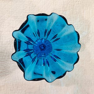 K&ouml;nnte beinhalten: Eine t&uuml;rkisfarbene Glasschale mit dunkelblauem Rand und einem bl&uuml;tenartigen Design in der Mitte. Die Schale hat einen ger&uuml;schten Rand und eine gl&auml;nzende Oberfl&auml;che, ideal f&uuml;r dekorative Zwecke oder zum Servieren. Durchmesser ca. 20 cm.