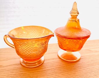 Könnte beinhalten: Ein orangefarbenes Sahne- und Zuckerdosen-Set aus Glas. Der Sahneaufgießer hat einen Griff und einen Ausguss, während die Zuckerdose einen dekorativen Deckel hat. Beide Teile haben ein geprägtes Blumenmuster und stehen auf einer Holzoberfläche.