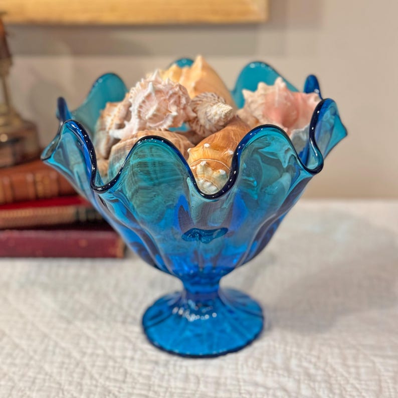 K&ouml;nnte beinhalten: Eine leuchtend blaue Glasschale mit gewelltem Rand, gef&uuml;llt mit verschiedenen Muscheln. Die Schale hat einen Sockel. Die Muscheln sind in verschiedenen Gr&ouml;&szlig;en und Farben gehalten und bilden ein maritimes Thema. Die Schale hat einen Durchmesser von ca. 20 cm.