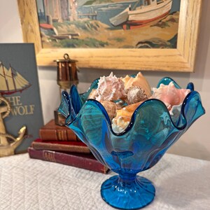 K&ouml;nnte beinhalten: Eine leuchtend blaue Glasschale mit gewelltem Rand, gef&uuml;llt mit verschiedenen Muscheln. Die Schale steht auf einem Sockel. Ein Gem&auml;lde einer K&uuml;stenlandschaft und B&uuml;cher befinden sich im Hintergrund.