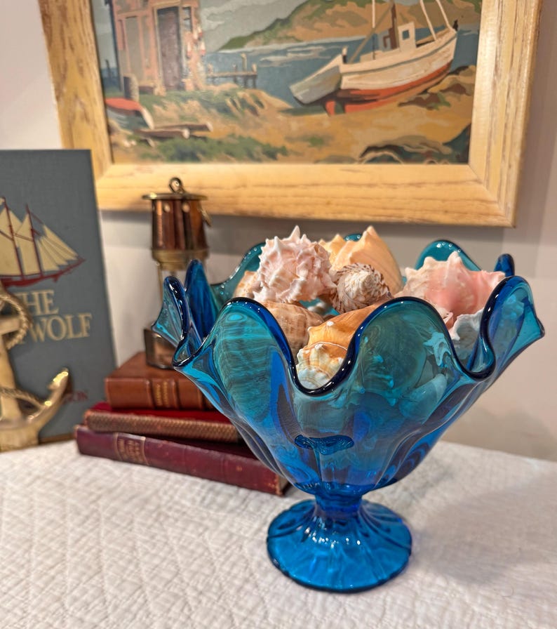 K&ouml;nnte beinhalten: Eine leuchtend blaue Glasschale mit gewelltem Rand, gef&uuml;llt mit verschiedenen Muscheln. Die Schale steht auf einem Sockel. Ein Gem&auml;lde einer K&uuml;stenlandschaft und B&uuml;cher befinden sich im Hintergrund.