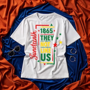May include: White t-shirt with the text "Juneteenth" in red script on the left side. The shirt features the text "1865 THEY NOT LIKE US" in green, yellow, and red, with star and triangle accents.