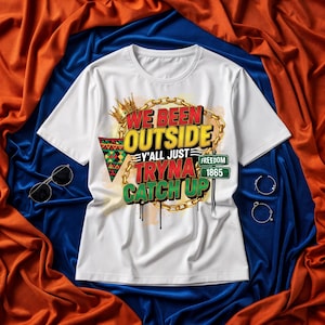 May include: White t-shirt featuring the text "WE BEEN OUTSIDE Y'ALL JUST TRYNA CATCH UP" in red, yellow, and green. The design includes a gold crown, chain, and a triangle with a colourful pattern. Sunglasses and earrings are also visible.