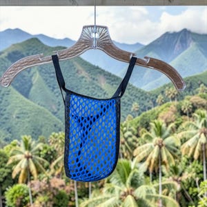 May include: A blue mesh bag with black trim and straps hangs from a clear plastic hanger. The bag has a diamond-shaped pattern. The background features a lush green landscape with mountains and palm trees.