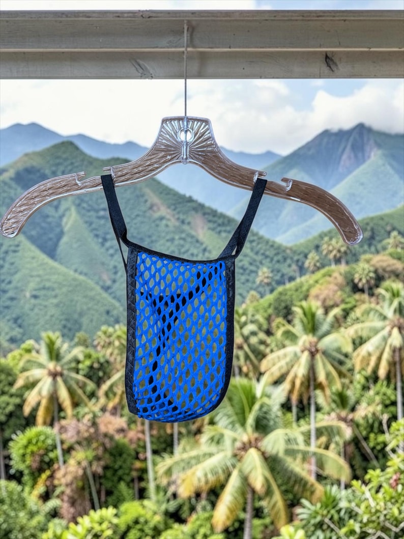 May include: A blue mesh bag with black trim and straps hangs from a clear plastic hanger. The bag has a diamond-shaped pattern. The background features a lush green landscape with mountains and palm trees.