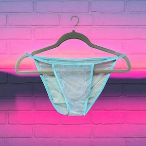 May include: Light blue underwear with a sheer, see-through design, trimmed with a matching light blue trim. The underwear is hanging on a gray hanger against a vibrant pink and purple brick wall.