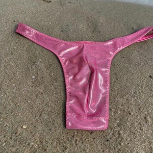 May include: A shiny pink thong swimsuit lies on a sandy beach. The swimsuit has a low-rise front and thin straps. The fabric has a subtle shimmer, catching the light. The background shows the edge of the ocean with small waves.