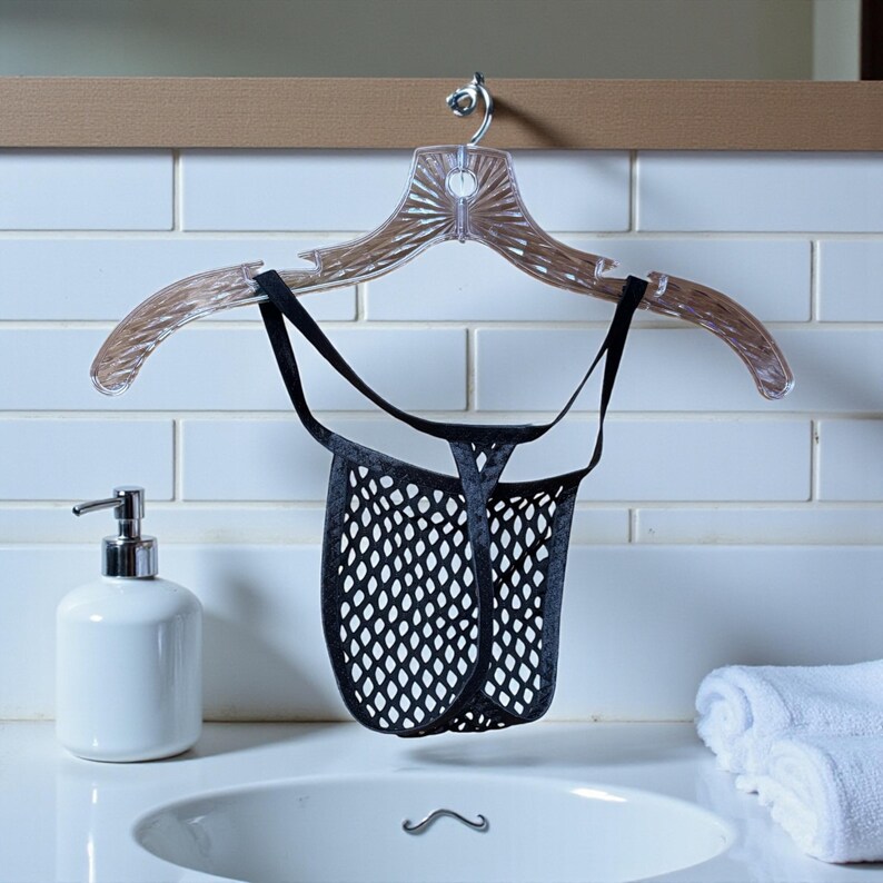 May include: Black mesh thong underwear hanging on a clear plastic hanger. The underwear is suspended above a white sink with a chrome faucet. A white soap dispenser and folded white towels are also visible.