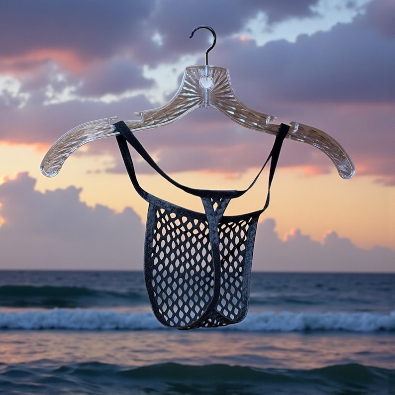 May include: A black mesh thong hangs from a clear plastic hanger against a backdrop of a sunset over the ocean. The thong has a diamond-shaped pattern and black straps. The hanger is clear and has a decorative design.