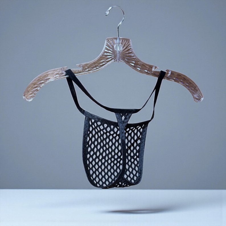 May include: A black mesh thong suspended from a clear plastic hanger. The thong has a net-like design and is held by thin black straps. The hanger is clear and has a decorative design.