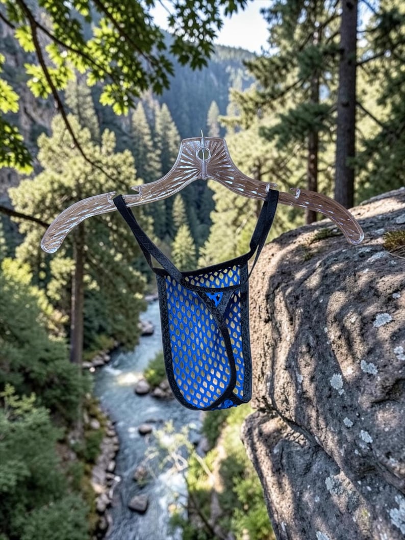 May include: A blue mesh pouch underwear with black trim, hanging from a clear plastic hanger. The background features a scenic view of a river and forest.