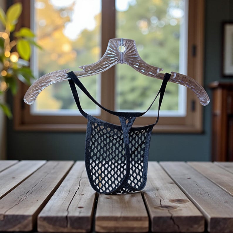 May include: A black mesh thong hangs from a clear plastic hanger. The thong has a diamond-shaped pattern and a thin waistband. The hanger is suspended against a blurred background of a window and wooden table.