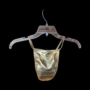 May include: A gold metallic thong swimsuit hangs from a clear plastic hanger against a black background. The swimsuit has thin straps and a shiny, textured appearance. The edges are neatly finished.