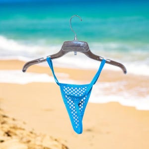 May include: A blue mesh bikini bottom hanging on a clear plastic hanger against a beach background.