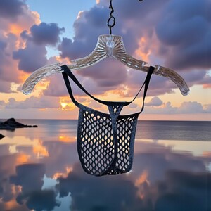 May include: A black mesh thong underwear hangs from a clear plastic hanger. The underwear has a unique design with a net-like pattern. The background features a sunset over the ocean, with the sky reflecting in the water.