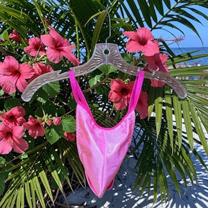 May include: A bright pink swimsuit hangs on a clear plastic hanger against a backdrop of tropical flowers and foliage. The swimsuit has a high-cut leg and a halter-style top. The background includes pink hibiscus flowers, green palm leaves, and a glimpse of the ocean.