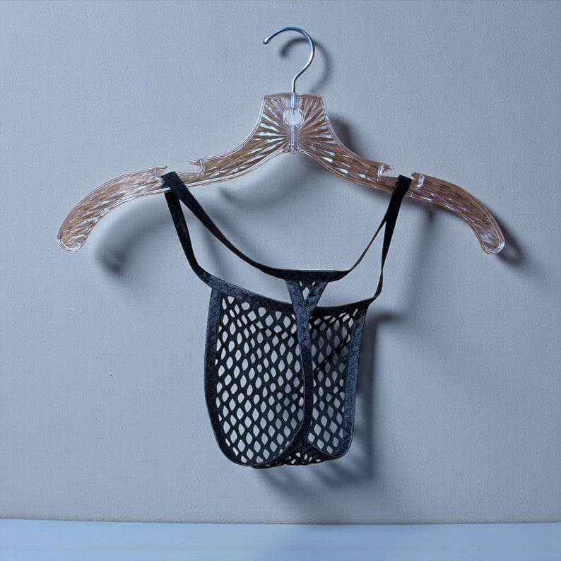 May include: A black mesh thong hangs from a clear plastic hanger. The thong has a low-rise design with a net-like pattern. The hanger is clear with a decorative design.