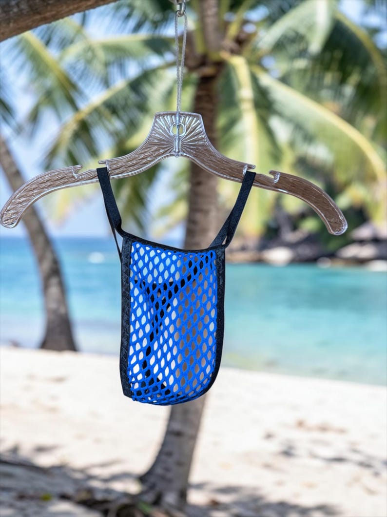 May include: A blue mesh bag with black trim hangs from a clear plastic hanger. The bag is suspended against a backdrop of a beach with palm trees and turquoise water.