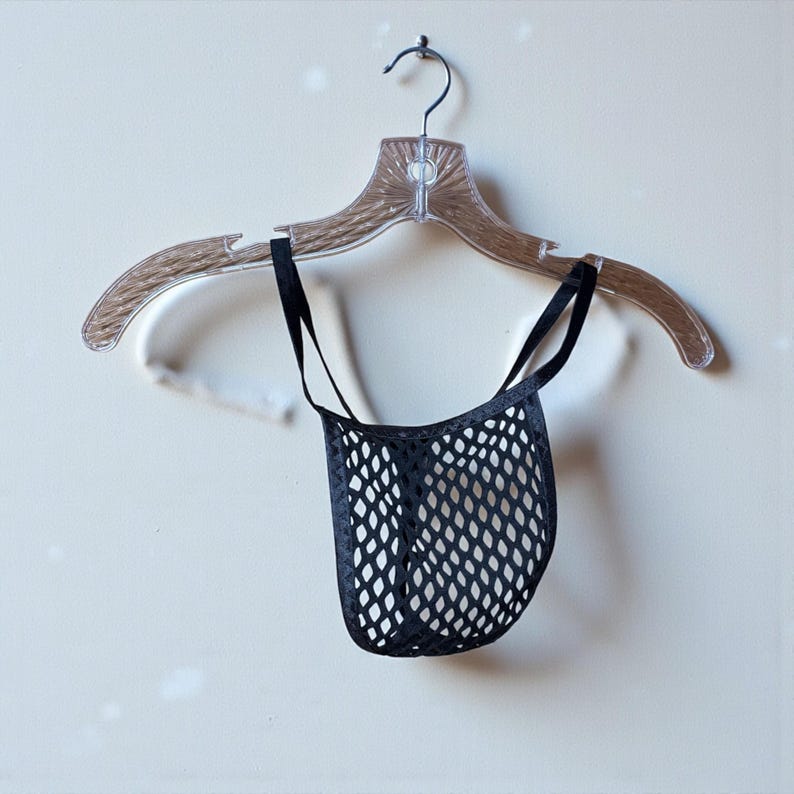 May include: A black mesh thong with thin black straps hangs from a clear plastic hanger. The thong has a diamond-shaped pattern. The hanger is suspended against a light-colored wall.