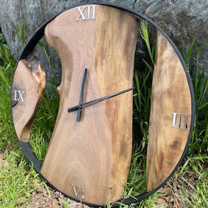 Może przedstawiać: Rustykalny drewniany zegar ścienny z naturalnym usłojeniem. Tarcza zegara posiada rzymskie cyfry i czarne wskazówki. Zegar jest zamknięty w czarnej metalowej ramie, o średnicy około 40 cm.