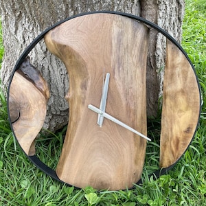 Op de afbeelding: Een houten klok met een natuurlijke houten afwerking en een zwart metalen frame. De wijzerplaat van de klok is gemaakt van een enkel stuk hout met een unieke vorm. De wijzers van de klok zijn zilver.