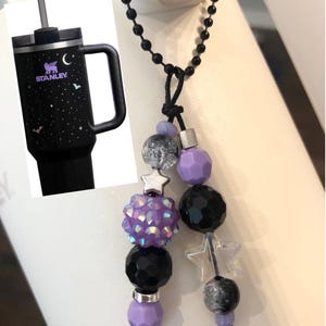 May include: Black Stanley tumbler with a handle, featuring a purple logo, moon, stars, and bats design. A beaded charm with purple, black, and clear beads, and star accents hangs from a black chain.