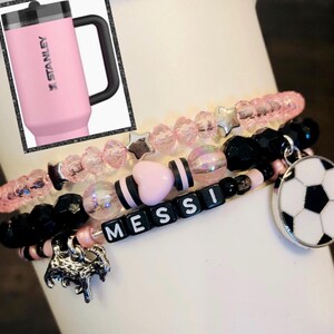May include: A set of four bracelets with pink and black beads, a pink heart charm, a silver star charm, a silver soccer ball charm, and black letter beads spelling "MESSI".