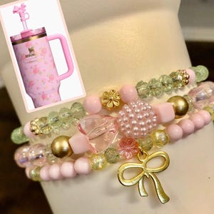May include: A stack of three beaded bracelets in shades of pink, green, and gold. The bracelets feature various beads, including pearls, faceted beads, and gold accents. A gold bow charm hangs from the bottom bracelet. A pink floral Stanley tumbler is in the background.