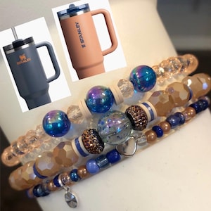 May include: Four layered bracelets with blue, peach, and silver beads. The bracelets feature a heart charm and a crystal charm. The bracelets are stacked on a white surface.