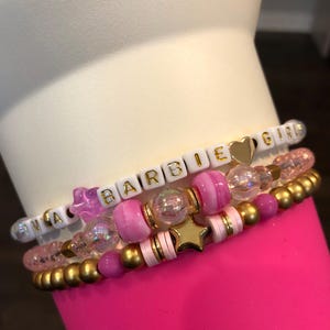 Pu&ograve; includere: Un set di quattro braccialetti con perline rosa, bianche e dorate. I braccialetti compongono la scritta "Barbie Girls" con perline a lettere bianche e lettere dorate. I braccialetti sono impilati uno sopra l'altro su una superficie rosa.