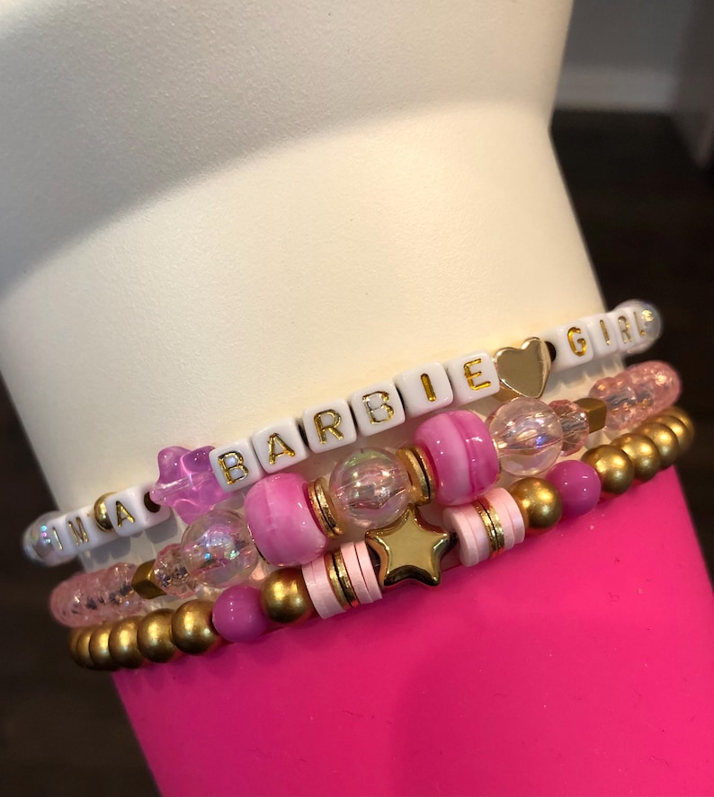 Pu&ograve; includere: Un set di quattro braccialetti con perline rosa, bianche e dorate. I braccialetti compongono la scritta "Barbie Girls" con perline a lettere bianche e lettere dorate. I braccialetti sono impilati uno sopra l'altro su una superficie rosa.