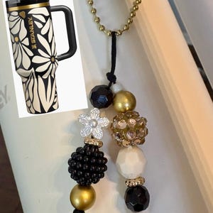 May include: A black and gold Stanley tumbler with a daisy pattern and a black handle. A beaded keychain with black, gold, and white beads and a flower charm hangs nearby. The tumbler is a popular item for carrying beverages.