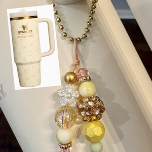 May include: A decorative charm featuring a gold-toned ball chain and a collection of beads in shades of yellow, gold, and clear crystal, including a flower-shaped bead. An inset image shows a cream-colored Stanley LoveShackFancy tumbler.