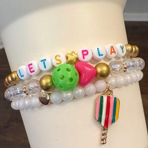 May include: A stack of beaded bracelets with a pickleball theme. The top bracelet spells "LET'S PLAY" with colorful letter beads. Other beads include gold, white, and iridescent beads, and a charm of a pickleball paddle.