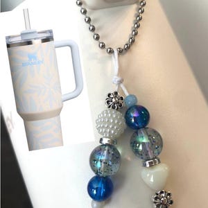 May include: A cream-colored Stanley tumbler with a light blue floral design and handle. A beaded keychain with blue, iridescent, and white beads, silver accents, and flower charms hangs nearby.